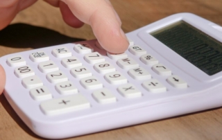 Homeowner sits at a clean, modern desk, using a calculator to review documents related to selling their home, with a set o...