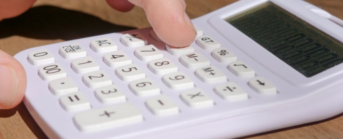 Homeowner sits at a clean, modern desk, using a calculator to review documents related to selling their home, with a set o...